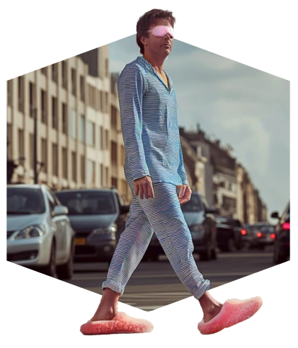Man loopt overdag op straat met pyjama, roze slippers en een nachtmasker over zijn ogen